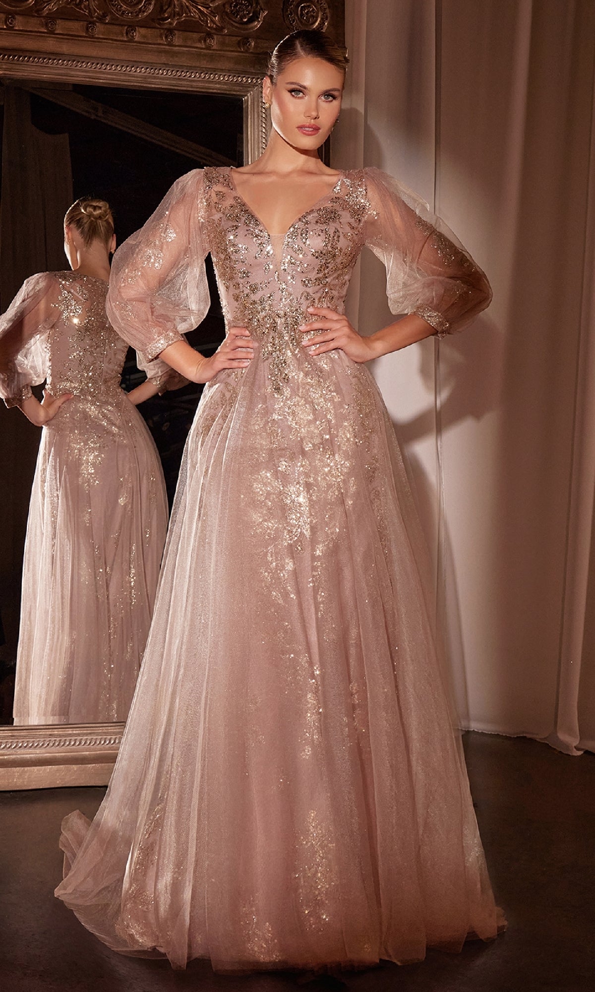 A woman wearing the Ladivine CD0102 Sheer-Sleeve Long Prom Dress in blush pink, featuring puff sleeves and silver embellishments, stands with hands on hips before a mirror.